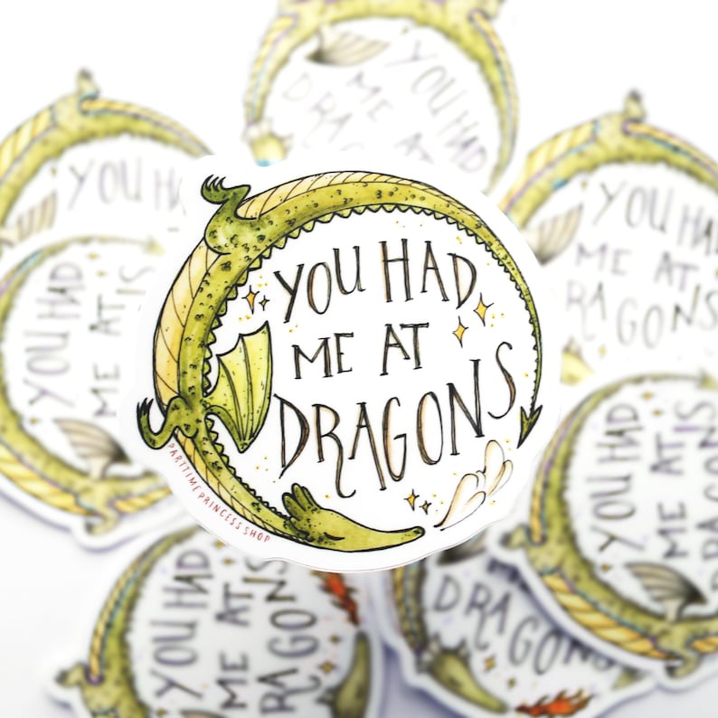 Op de afbeelding: Een ronde sticker met een groene draak die om de tekst "You had me at dragons" gekruld is. De sticker heeft een witte achtergrond met gouden glitteraccenten.