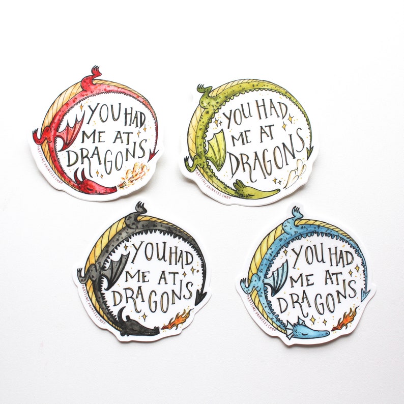 Op de afbeelding: Vier ronde stickers met een drakenontwerp. Elke sticker heeft een draak in een andere kleur met de tekst "You had me at dragons".