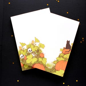 Peut inclure: Deux cartes blanches avec une illustration sur le thème d'Halloween. L'œuvre présente des citrouilles oranges, des feuilles vertes, des crânes et un chat noir. Le design est sur un fond noir avec des confettis en forme d'étoiles dorées.