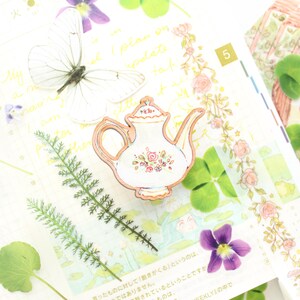 May include: A wooden teapot-shaped embellishment with a white body and a floral design. The teapot has a light brown outline and is surrounded by other decorative elements, including a white butterfly, green leaves, and purple flowers.