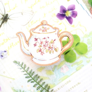 May include: A wooden brooch in the shape of a teapot, featuring a white body and a light brown border. The teapot is adorned with pink and yellow floral patterns. The brooch is set against a background with a butterfly, flowers, and green leaves.