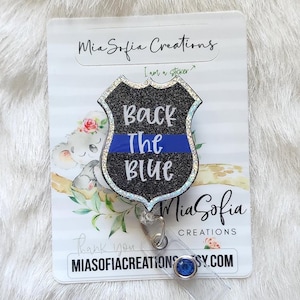May include: A badge-shaped retractable ID holder with the text "Back The Blue" in white and blue glitter. The badge is black with a silver glitter border and a blue line. The holder is attached to a white card with floral accents.
