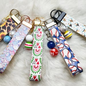 May include: A collection of six wristlet keychains with various patterns. Designs include leopard print with sunflowers, mermaid scales, watermelon slices, baseballs, and bees. Each keychain has a gold or black metal clasp and decorative beads.