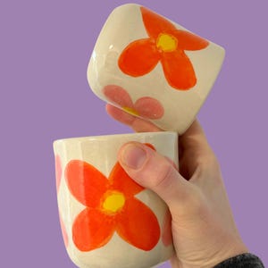 May include: Two ceramic mugs with a white base and a vibrant orange flower design with yellow centers. The mugs have a rounded shape and are held against a purple background. The flowers are painted in a bold, playful style.
