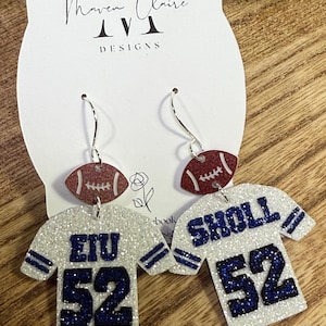 May include: A pair of dangle earrings featuring a glittery white football jersey shape with dark blue text reading EIU 52 and SHOLL 52, accented by a small glittery maroon football charm at the top, suspended from silver-toned metal earring hooks.