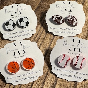 May include: Four pairs of sport-themed earrings on white cards with the text "Maven Claire Designs" and a website address. The earrings feature a soccer ball, football, basketball, and baseball.