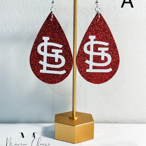May include: Red glitter teardrop earrings with a white St. Louis Cardinals logo. The earrings are hanging from a gold stand.