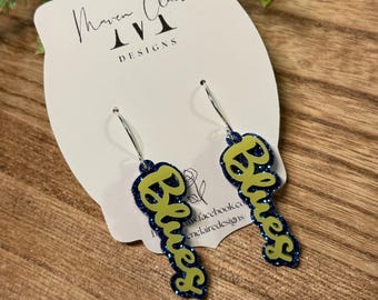 May include: A pair of earrings with the word "Blues" in a neon green script font, outlined in dark blue glitter. The earrings hang from silver hoops. The earrings are displayed on a white card with the text "Maven Claire DESIGNS".