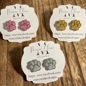 May include: Three pairs of glitter paw print earrings on white cards. The cards have the text "Maven Claire Designs" and a website address. The earrings are pink, gold, and silver.
