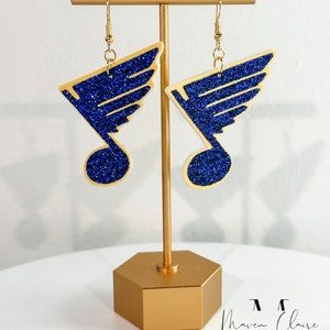 May include: A pair of blue and gold glitter earrings with a musical note and wing design. The earrings are displayed on a gold stand.