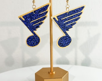 May include: A pair of blue and gold glitter earrings with a musical note and wing design. The earrings are displayed on a gold stand.