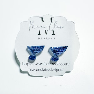 May include: A pair of blue glitter earrings featuring the St. Louis Blues hockey team logo. The earrings are on a white card with the text "Maven Claire Designs" and a website address.