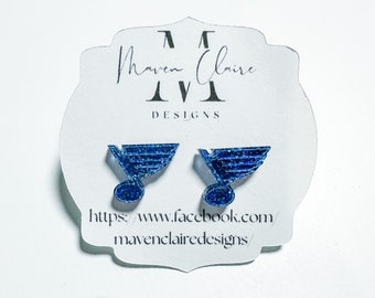 May include: A pair of blue glitter earrings featuring the St. Louis Blues hockey team logo. The earrings are on a white card with the text "Maven Claire Designs" and a website address.