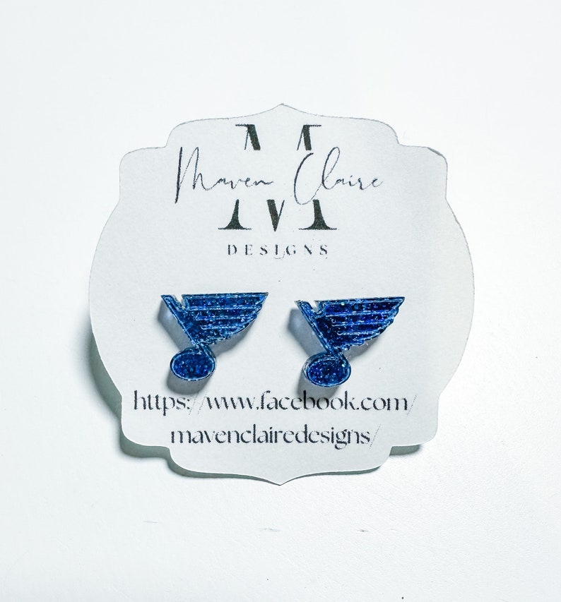May include: A pair of blue glitter earrings featuring the St. Louis Blues hockey team logo. The earrings are on a white card with the text "Maven Claire Designs" and a website address.