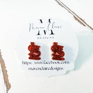 May include: A pair of red glitter dollar sign earrings on a white card with the text "Maven Claire Designs" and a website address.