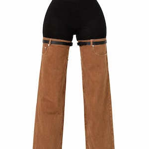 May include: Black shorts with brown denim chaps. The chaps are attached to a black belt with a buckle. The denim chaps have front pockets and a wide leg design. The background is white.
