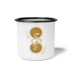 Può includere: Tazza in smalto bianco con bordo nero che presenta un design a serpente dorato con un motivo circolare. Il serpente è avvolto attorno a due cerchi sovrapposti con un motivo a punti.