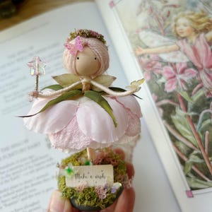 May include: A handcrafted fairy doll with a pink petal skirt, holding a star wand. The doll has a wooden head, pink yarn hair, and a butterfly hair accessory. The base is decorated with moss and a "Make a wish" sign. A book with illustrations is in the background.