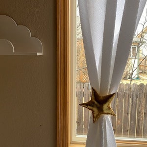 Könnte beinhalten: Ein weißer, durchsichtiger Vorhang mit einem goldenen, sternförmigen Vorhangbinder. Der Vorhang hängt an einem Fenster mit Blick auf einen Garten.