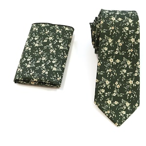 Op de afbeelding: Een donker bosgroene das en een bijpassend gevouwen pochet, beide voorzien van een herhalend patroon van kleine, delicate witte bloemetjes met dunne groene steeltjes en blaadjes verspreid over het oppervlak van de stof.