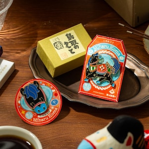 Może przedstawiać: Kolekcja japońskich pamiątek z motywem konia. Zawiera okrągłą przypinkę, prostokątny amulet i małe pudełko, wszystkie z kolorowymi wzorami. Przypinka i amulet mają napis "Miharu Koma".