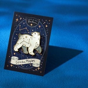 Puede incluir: Un pin de esmalte de color dorado con un oso polar blanco con un diseño de constelación. El pin está en una tarjeta negra con estrellas doradas y el texto "The Star Drifters" y "Ursa Major".