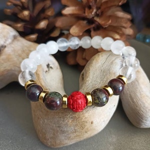 May include: A white beaded bracelet with a red and green stone bead and gold accents.
