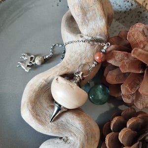 Peut inclure: Un pendule en bois avec une chaîne argentée et un charme en pierre rouge. Le pendule repose sur un morceau de bois flotté avec des pommes de pin et des perles de verre autour.