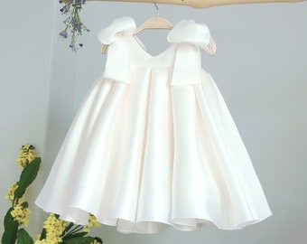 May include: A white satin dress with large bows on the shoulders. The dress is hanging on a wooden hanger.
