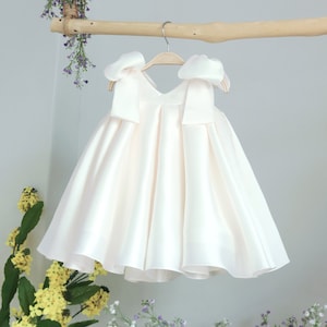 Könnte beinhalten: Ein weißes Satin-Kleid mit großen Schleifen an den Schultern. Das Kleid hängt an einem Holzbügel.