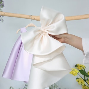 Può includere: Un abito di raso lilla chiaro con un grande fiocco di raso avorio su un appendiabiti in legno.