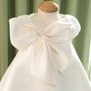 Könnte beinhalten: Weißes Satinkleid mit großer Schleife am Ausschnitt. Die Schleife ist ein Blickfang und verleiht dem Kleid einen Hauch von Eleganz. Das Kleid wird auf einer Schaufensterpuppe präsentiert.