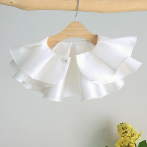 Puede incluir: Collar blanco con volantes y cierre de botón. El collar está colgado en una percha de madera.