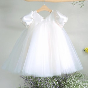 May include: A white baby dress with a tulle skirt and satin bodice, hanging from a wooden hanger. The dress has short, puffed sleeves and a V-neckline. The dress is displayed against a soft gray background, with a bouquet of flowers at the bottom.