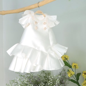 May include: A white satin dress with a ruffled skirt and sleeves. The dress has three pearl-like buttons on the front.