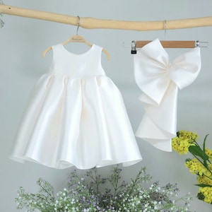 Könnte beinhalten: Ein weißes Satin-Kleid mit einem ärmellosen Oberteil und einem vollen Rock. Das Kleid hängt an einem Holzbügel. Eine passende weiße Satinschleife hängt ebenfalls an einem Holzbügel.