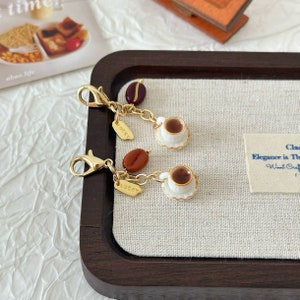 Könnte beinhalten: Ein goldenes Charm-Armband mit zwei Miniatur-Kaffeetassen, zwei Kaffeebohnen und zwei goldenen Charms mit der Aufschrift "Lucky". Das Armband liegt auf einem Tablett aus Holz mit einer weißen Stoffauskleidung.