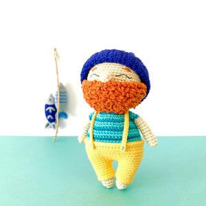 May include: A crocheted toy fisherman with a blue hat, orange beard, blue and green striped shirt, and yellow pants. The toy is holding a fishing rod with a blue fish.