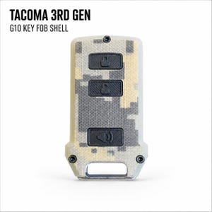 May include: A G10 key fob shell for a 3rd Gen Tacoma featuring a tan and gray digital camouflage pattern. The rectangular shell has three black rubberized buttons with lock, unlock, and panic icons, secured by two small black screws at the base.