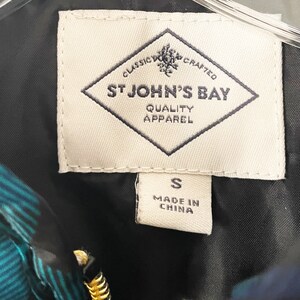 May include: Close-up of a tag from a garment. The tag is cream-colored with the words "CLASSIC CRAFTED ST JOHN'S BAY QUALITY APPAREL" in navy blue. Below the main tag is a smaller tag with the letter "S" and "MADE IN CHINA".