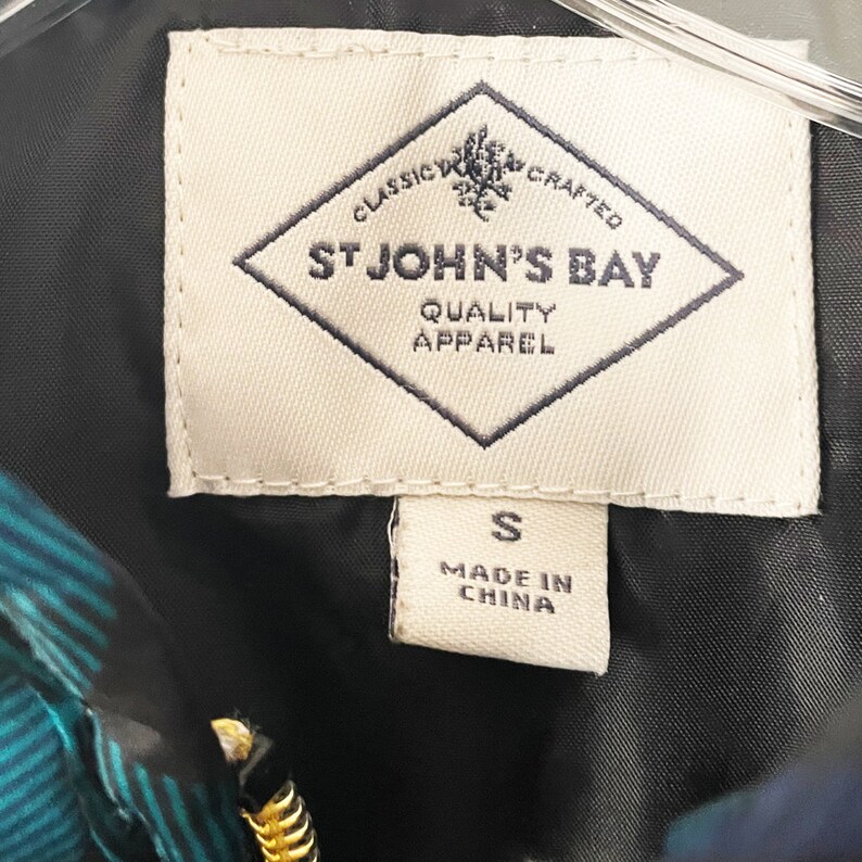 May include: Close-up of a tag from a garment. The tag is cream-colored with the words "CLASSIC CRAFTED ST JOHN'S BAY QUALITY APPAREL" in navy blue. Below the main tag is a smaller tag with the letter "S" and "MADE IN CHINA".