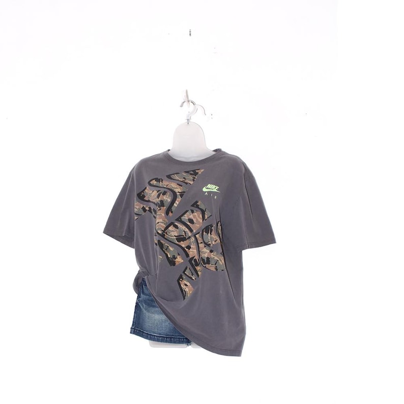Pode incluir: Camiseta cinza Nike com design de camuflagem e o logotipo Nike Air em verde. A camiseta &eacute; combinada com uma saia jeans. A camiseta tem mangas curtas e gola redonda.