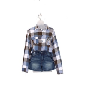 May include: A blue, brown, and white plaid cropped flannel shirt paired with denim jean shorts. The shirt has two chest pockets and a button-down front. The shorts are a dark wash denim.