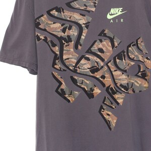 Pode incluir: Uma camiseta cinza com um grande design abstrato em tons de marrom, verde e preto. O logotipo da Nike e "AIR" s&atilde;o impressos em verde neon. A camiseta tem mangas curtas e gola redonda.