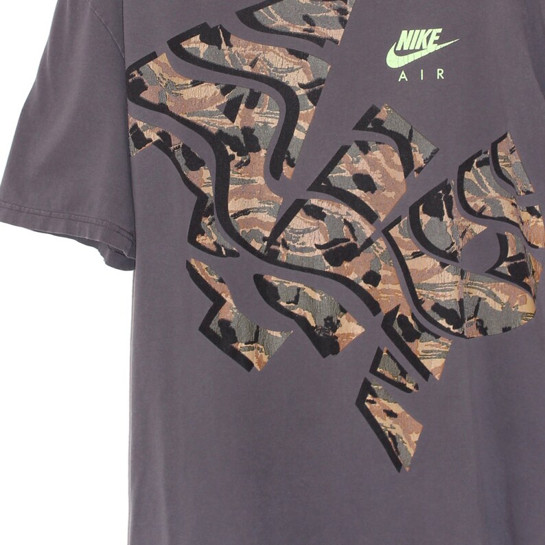 Pode incluir: Uma camiseta cinza com um grande design abstrato em tons de marrom, verde e preto. O logotipo da Nike e "AIR" s&atilde;o impressos em verde neon. A camiseta tem mangas curtas e gola redonda.