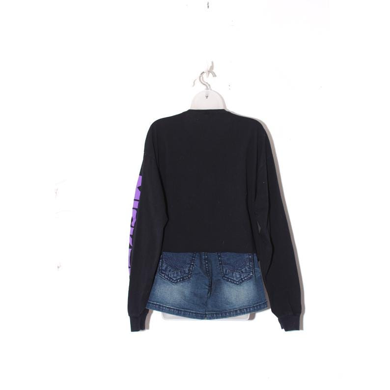 Op de afbeelding: Zwart shirt met lange mouwen met een aangehechte spijkerbroekrok. Het shirt heeft paarse tekst op de linkermouw. De spijkerbroekrok is een medium wassing met zichtbare stiksels en zakken. Het shirt hangt aan een witte hanger.