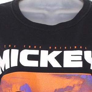 Op de afbeelding: Zwart T-shirt met ronde hals met de tekst "THE TRUE ORIGINAL MICKEY" in wit en oranje. Het shirt heeft een kleurrijke afbeelding van een gezicht. Een klassieke stijl.