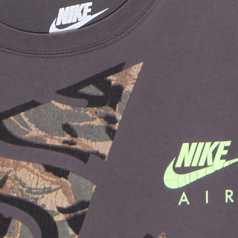 Pode incluir: Close-up de uma camiseta Nike cinza escuro. A camisa apresenta um design de camuflagem em tons de marrom, verde e preto. O logotipo da Nike &eacute; vis&iacute;vel em branco no topo e em verde neon com "AIR" impresso abaixo.