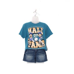 May include: Teal Arizona Diamondbacks Hall of Fame t-shirt with a graphic of two baseball players and the text "Hall of Fame". Paired with denim jean shorts. The shirt is displayed on a white mannequin.