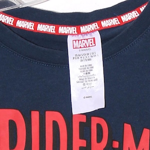 以下が含まれることがあります： 襟に赤いMARVELの文字が入ったネイビーブルーのTシャツ。MARVELのロゴとテキストが入った白いタグが付いています。シャツの下部には大きな赤い「DIDER-M」グラフィックがあります。